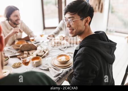 Young joyful asian man with dark hair in eyeglasses and hoodie sitting at the table happily looking aside. Group of international friends having lunch Stock Photo