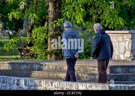 Gentlemen, two older men are walking through an open-air park at the time of the infectious Covid - 19. They use masks and gloves. Stock Photo