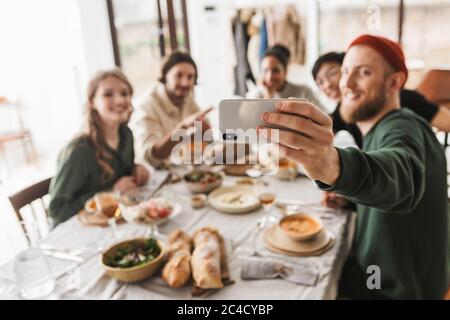 Group of attractive international friends sitting at the table full of food spending time in cozy cafe. Young colleagues happily taking photo on Stock Photo