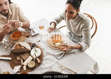 Top view of smiling african american woman sitting at the table eating cream soup with slice of bread in hand. Group of young international friends Stock Photo