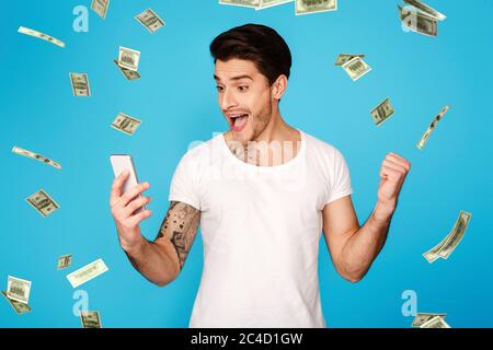Lucky Winner. Excited Man With Smartphone Screaming With Joy Under Money Shower Stock Photo