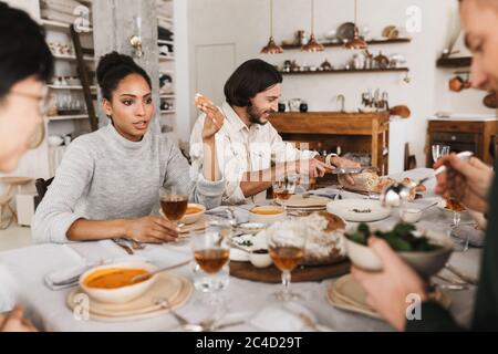 Pretty afro american woman sitting at the table thoughtfully talking while man near happily cutting bread. Group of young international friends Stock Photo
