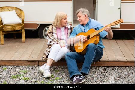 Camping fun. Romantic senior husband singing to his charming wife near their mobile home Stock Photo