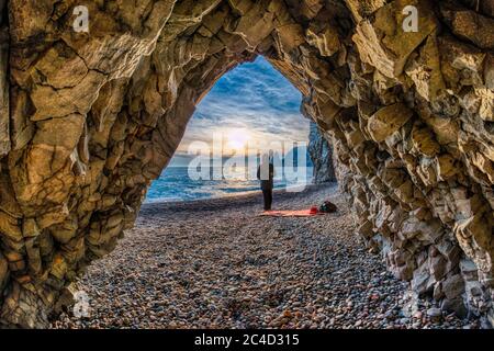 view from the stone cave on the sunset sea and the beach, the volcanic rock of the cave is lit by the warm setting sun. The concept of calmness Stock Photo