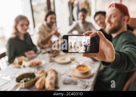 Group of attractive international friends sitting at the table with food spending time together. Young colleagues joyfully taking photo on modern Stock Photo