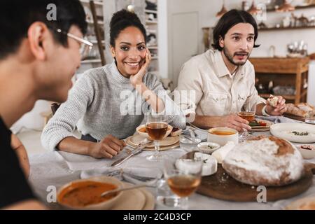 Pretty smiling african american woman with dark curly hair sitting at the table leaning on hand happily looking at friend. Group of young Stock Photo