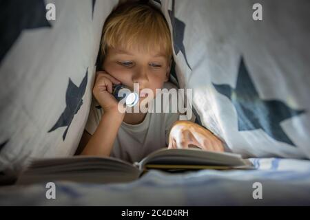 A little boy reads a book with a flashlight under the covers at night. Stock Photo