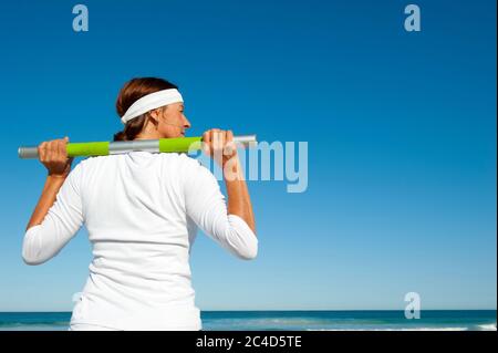 Attractive and active senior woman relaxed standing with weight over her shoulder looking over ocean after healthy exercise, isolated with blue sky as Stock Photo