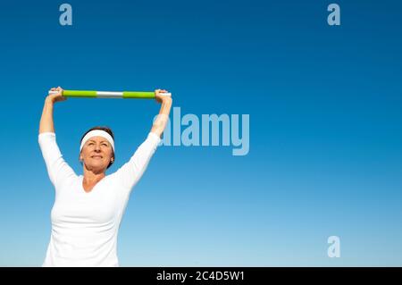 Attractive and active senior woman relaxed standing with open eyes, weight over her head doing healthy exercise, isolated with blue sky as background Stock Photo