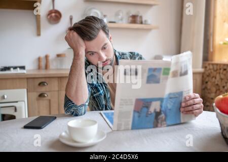 Man in a checkered shirt sitting at the table, reading newspaper and looking disturbed Stock Photo