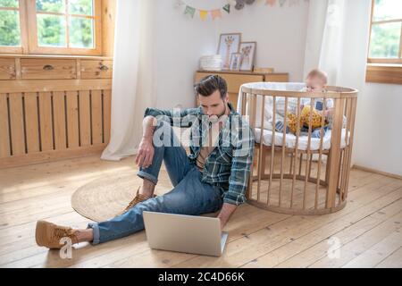 Dark-haired father sitting on the floor, working on a laptop while his little son playing Stock Photo