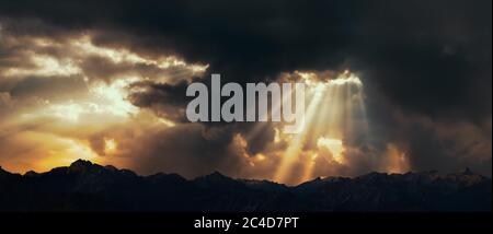 Rays of light shining through dark clouds over mountains. Cinematic scene. Stock Photo