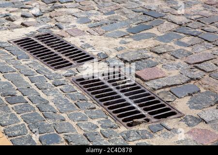 the 2 iron grates of the drainage system hatch on the road paved with paving stones, closeup of a sewer grates. Stock Photo
