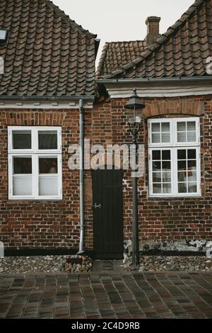 Narrow wooden door between old brick houses with white framed windows with the street lamp and stone road in front of them Stock Photo