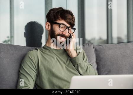 Image of joyful bearded man talking on smartphone and using laptop while sitting in living room Stock Photo