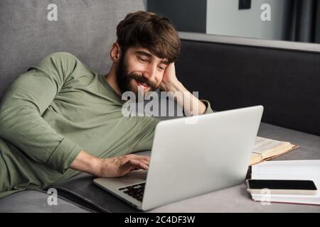 Image of joyful young man in casual wear using laptop and smiling while lying on sofa in living room Stock Photo