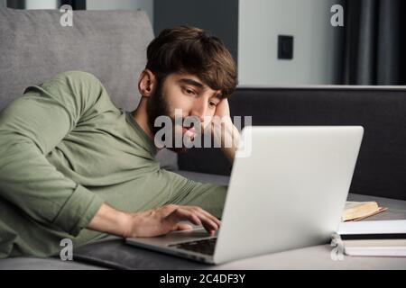 Image of concentrated young man in casual wear using laptop while lying on sofa in living room Stock Photo