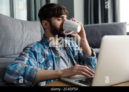 Image of handsome bearded man drinking coffee and using notebook while sitting on floor in living room Stock Photo