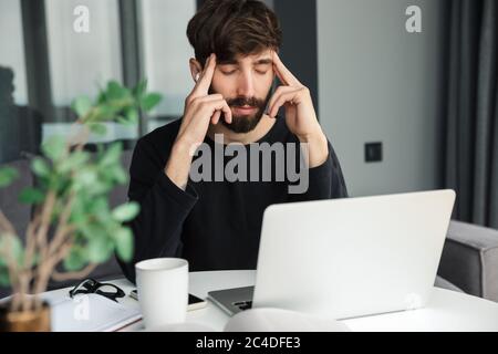 Image of tired man with headache using wireless earphones and laptop while working in living room Stock Photo