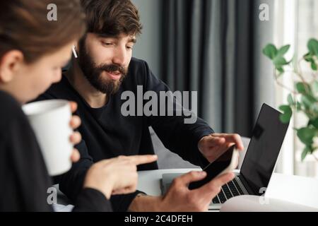 Image of young pleased colleagues discussing project and using cellphone while working at table in cozy room