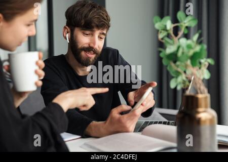 Image of young joyful colleagues discussing project and using cellphone while working at table in cozy room