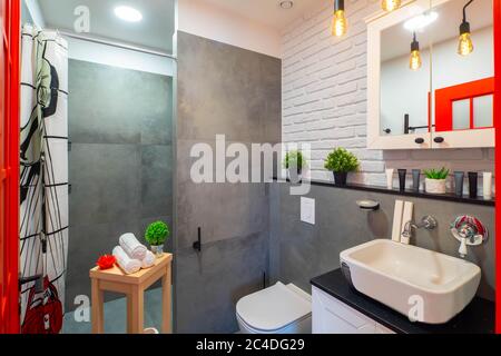Modern loft interior of bathroom. White sink and toilet. Brick wall. Grey tile. Red door. Stock Photo