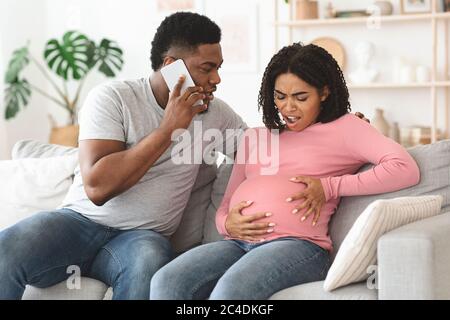 Pregnant black woman suffering, husband calling doctor, home interior Stock Photo