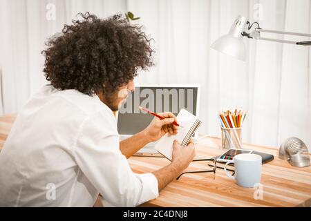 Side view of shaggy man making notes in his paper notebook or scratchpad sitting at wooden table with computer and cup of hot beverage on it. Memory Stock Photo