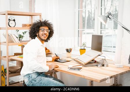 Intelligent freelanser smiles at camera sitting at home workplace. Creative young businessman working from home. Distant work or remote work concept Stock Photo