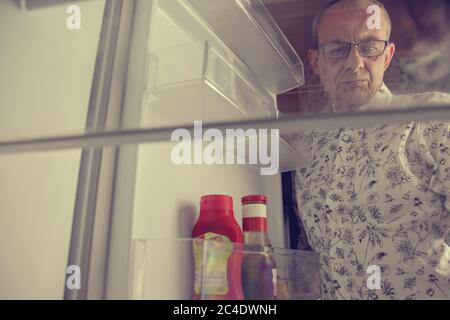 Senior man opening a refrigerator with little food. Selective focus. Hungry man looking for a late night meal in an empty fridge Stock Photo