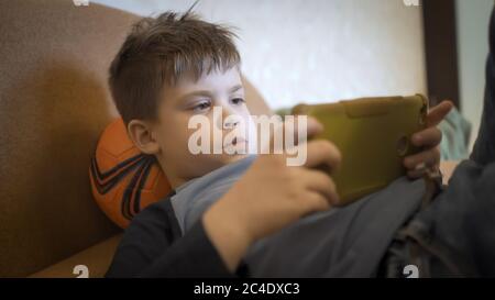 Serious boy playing games on mobile phone while lying on the couch with a ball under his head. Quarantine concept Stock Photo