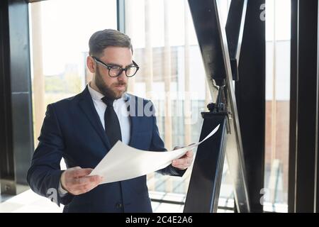 Bearded businessman in eyeglasses and in blue suit examining the business contract while standing at office Stock Photo