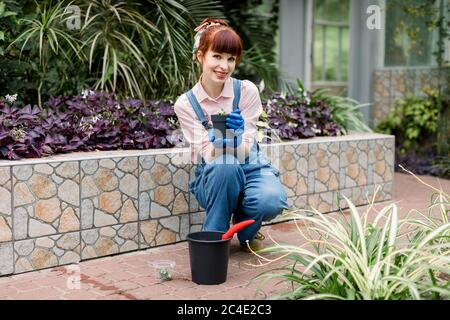 Attractive gardener woman in jeans overalls and rubber gloves, planting flowers and succulents in greenhouse, holding pot with soil in hands Stock Photo