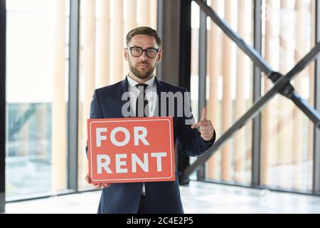 Portrait of real estate agent in suit pointing at placard in his hands and looking at camera he standing at office Stock Photo