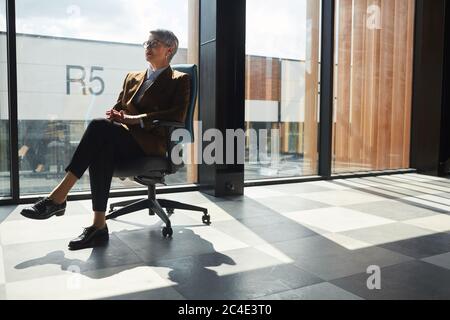Mature female leader sitting on chair and thinking about something against the big office window Stock Photo