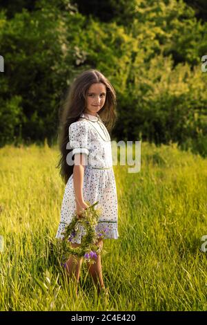 the girl is standing in the green grass, she made a wreath of flowers. Stock Photo