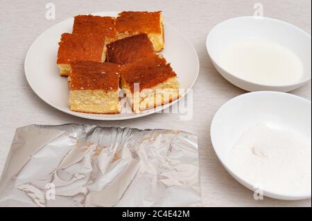 Traditional Brazilian dessert (known as 'Bolo Gelado') - Making step by step: Sliced cake, bowl with milk, grated coconut and aluminum foil. Stock Photo