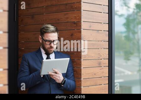Serious mature businessman in blue suit concentrating on his online work on digital tablet he standing outdoors Stock Photo