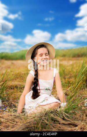 a girl with long hair is sitting on the hay in a straw hat Stock Photo