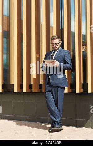 Mature businessman in blue formal suit standing outdoors near the office building and using tablet pc Stock Photo