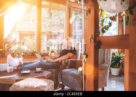 Man dressed black T-shirt and jeans sitting in a comfortable armchair, using a modern slim laptop and drinking tea in house sunroom living room. Dista Stock Photo