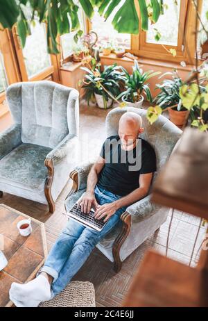 Man dressed black T-shirt and jeans sitting in a comfortable armchair, using a modern slim laptop and drinking tea in house sunroom living room. Dista Stock Photo