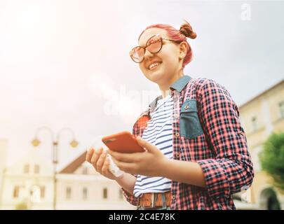 Portrait of smiling beautiful modern young female teenager with extraordinary hairstyle in checkered shirt holding a slim smartphone in hand. Modern t Stock Photo