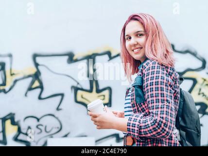 Portrait of cheerfully smiling Beautiful modern young female teenager with extraordinary hairstyle color in checkered shirt holding 'coffee to go' cap Stock Photo