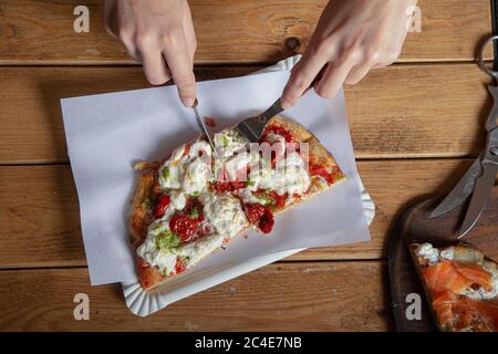 Woman's hands cut a piece of pizza on a wooden table. Italian pizza is eaten with a fork and knife. Foreground Stock Photo