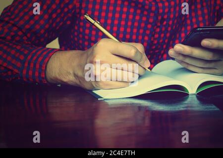 Young man writing on notebook about cost and holing phone using calculator in home office. Stock Photo
