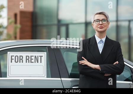 Portrait of mature businesswoman in black suit standing with arms crossed and looking at camera she suggesting the car for rent Stock Photo
