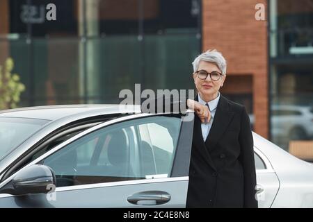 Portrait of mature businesswoman in eyeglasses standing near the car and looking at camera outdoors Stock Photo