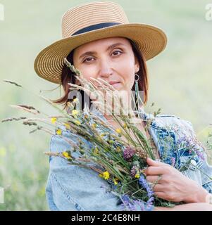 Calm portrait of a young woman with wildflowers bouquet. She dressed a jeans jacket, straw hat and light summer dress. Natural people's beauty concept