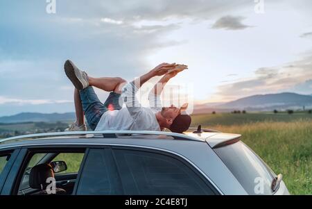 Middle-aged Man lying on the car roof and reading the paper bestseller book.He stopped his auto on the meadow with a beautiful valley view before the Stock Photo